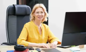 Uśmiechnięta wicedyrektor w żółtej bluzce siedzi przy biurku, przed nią komputer i słuchawki.