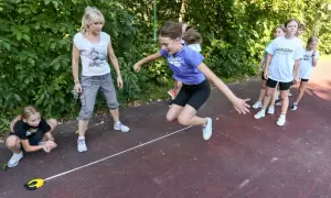 Dziewczynka skacze przez linkę podczas lekcji wychowania fizycznego w szkole. Nauczycielka i inne dzieci obserwują.