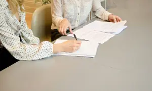 Dwie kobiety przy stole analizują dokumenty. Jedna z nich pisze coś w notatniku, być może wypełnia kartę rezydenta.