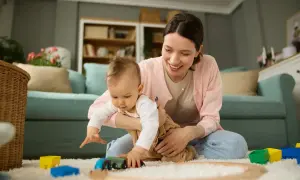 Aktywni rodzice w pracy to nie tylko biuro. Uśmiechnięta mama bawi się z dzieckiem klockami i pociągiem na dywanie.