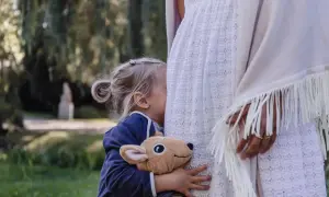 Małe, blond dziecko przytula pluszowego jelonka, chowając się za białą sukienką. To **wysoko wrażliwe dziecko** szuka ukojenia w ramionach mamy.