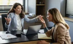 Dwie kobiety w biurze, jedna z wyrzutem gestykuluje, druga słucha. Scena może ilustrować, czym jest mobbing.