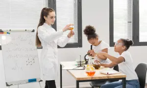 Nauczycielka demonstruje doświadczenie chemiczne, podczas gdy uczniowie pracują z mikroskopem i probówkami. Na tablicy widać wzory chemiczne, pomocne przy konkursy przedmiotowe arkusze.