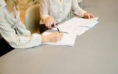 Dwie kobiety przy stole analizują dokumenty. Jedna z nich pisze coś w notatniku, być może wypełnia kartę rezydenta.