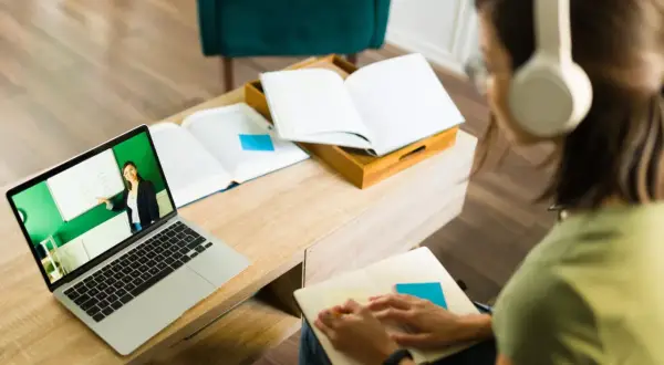 Dziewczyna w słuchawkach uczy się w domu. Na ekranie laptopa widać nauczycielkę prowadzącą lekcję w ramach polskiej szkoły internetowej.