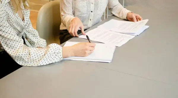 Dwie kobiety przy stole analizują dokumenty. Jedna z nich pisze coś w notatniku, być może wypełnia kartę rezydenta.