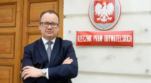 Mężczyzna w garniturze stoi przed budynkiem z godłem Polski i tabliczką "Rzecznik Praw Obywatelskich".