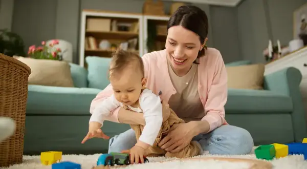 Aktywni rodzice w pracy to nie tylko biuro. Uśmiechnięta mama bawi się z dzieckiem klockami i pociągiem na dywanie.
