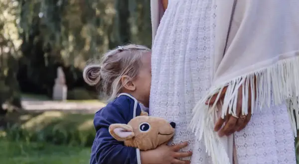 Małe, blond dziecko przytula pluszowego jelonka, chowając się za białą sukienką. To **wysoko wrażliwe dziecko** szuka ukojenia w ramionach mamy.