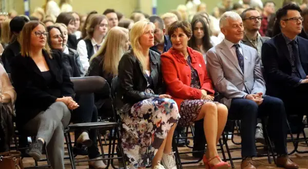 Uczestnicy siedzą na krzesłach, słuchając uważnie. Wśród nich widać osoby, które mogą być na radzie pedagogicznej, w tym kobiety w eleganckich strojach i mężczyzn w garniturach.
