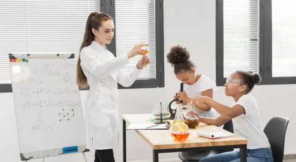 Nauczycielka demonstruje doświadczenie chemiczne, podczas gdy uczniowie pracują z mikroskopem i probówkami. Na tablicy widać wzory chemiczne, pomocne przy konkursy przedmiotowe arkusze.