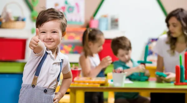 Uśmiechnięty chłopiec pokazuje kciuk w górę w przedszkolu, gdzie od kiedy zerówka jest obowiązkowa, dzieci bawią się i uczą.