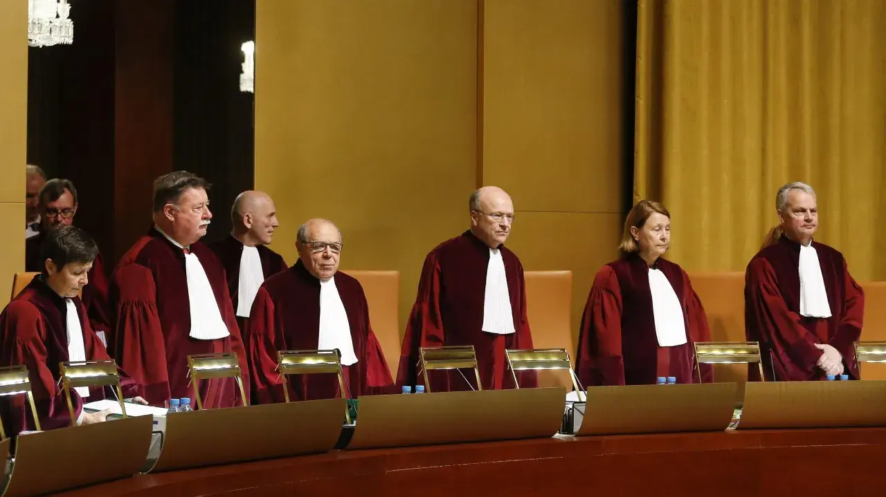Siedmiu sędziów w togach, w tym jedna kobieta, stoi w sali sądowej. Wszyscy mają na sobie białe żaboty. W tle widać złote zasłony.