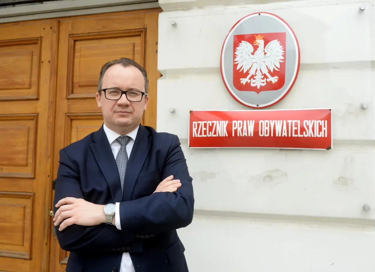 Mężczyzna w garniturze stoi przed budynkiem z godłem Polski i tabliczką "Rzecznik Praw Obywatelskich".