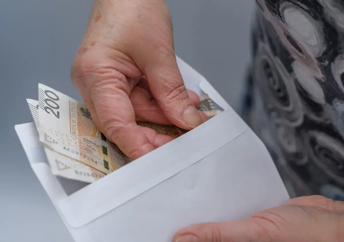Dłoń wkłada banknoty 200 zł do koperty. Ilustracja pokazuje przykłady przeliczenia emerytury kapitałowej na emeryturę powszechną.