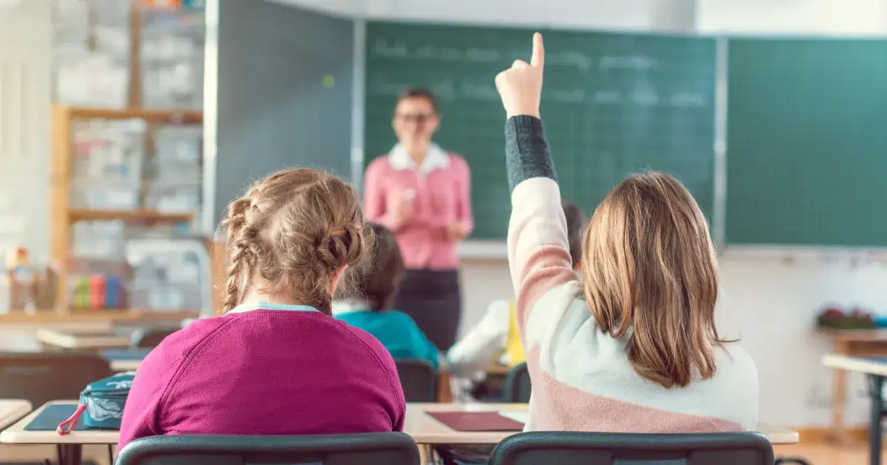 Uczniowie na lekcji, jedna dziewczynka podnosi rękę. Nauczycielka wyjaśnia materiał. Ciekawe, ile zarabia nauczyciel za taką pracę?