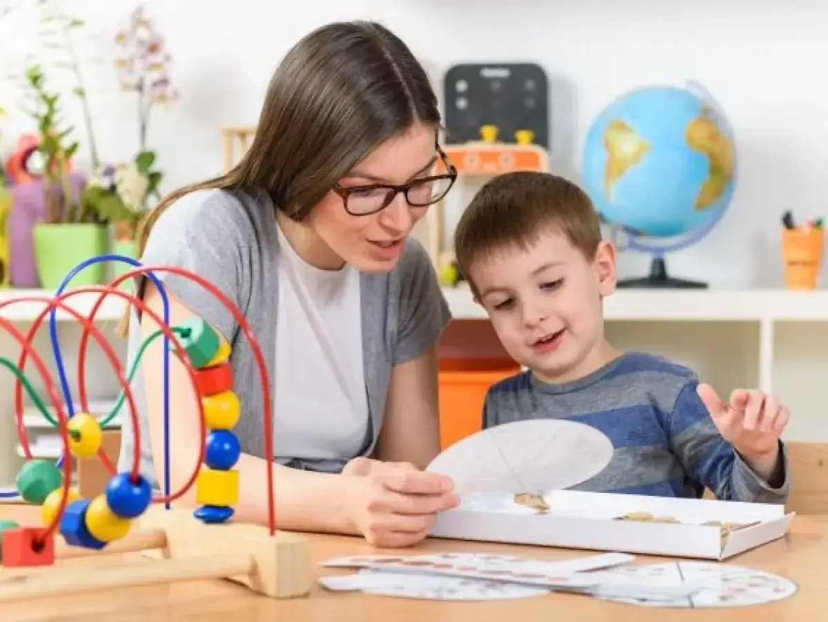 Pomoc psychologiczno pedagogiczna przykłady: nauczycielka z okularami i chłopiec bawią się koralikami i układanką.