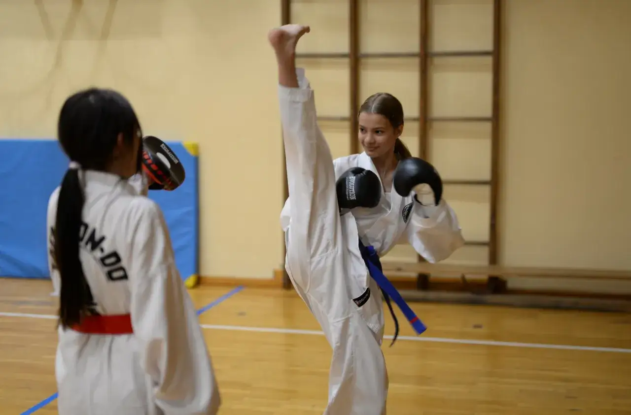 Dziewczynka w białym stroju do taekwondo wykonuje wysokie kopnięcie. To świetny przykład wychowania fizycznego w szkołach, rozwijający sprawność i pewność siebie.