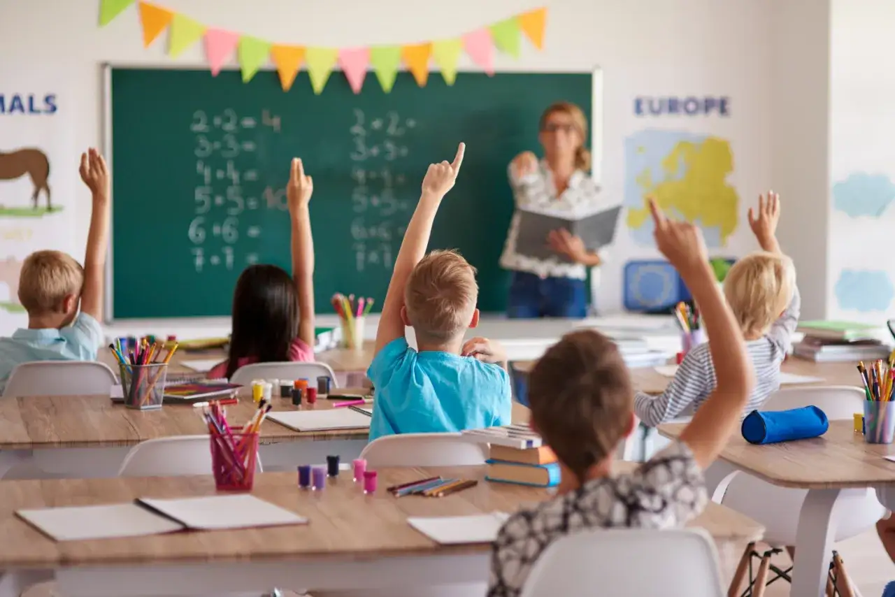 Dzieci na lekcji matematyki podnoszą ręce, by odpowiedzieć na pytanie nauczycielki. Obowiązek szkolny trwa do 18 roku życia.