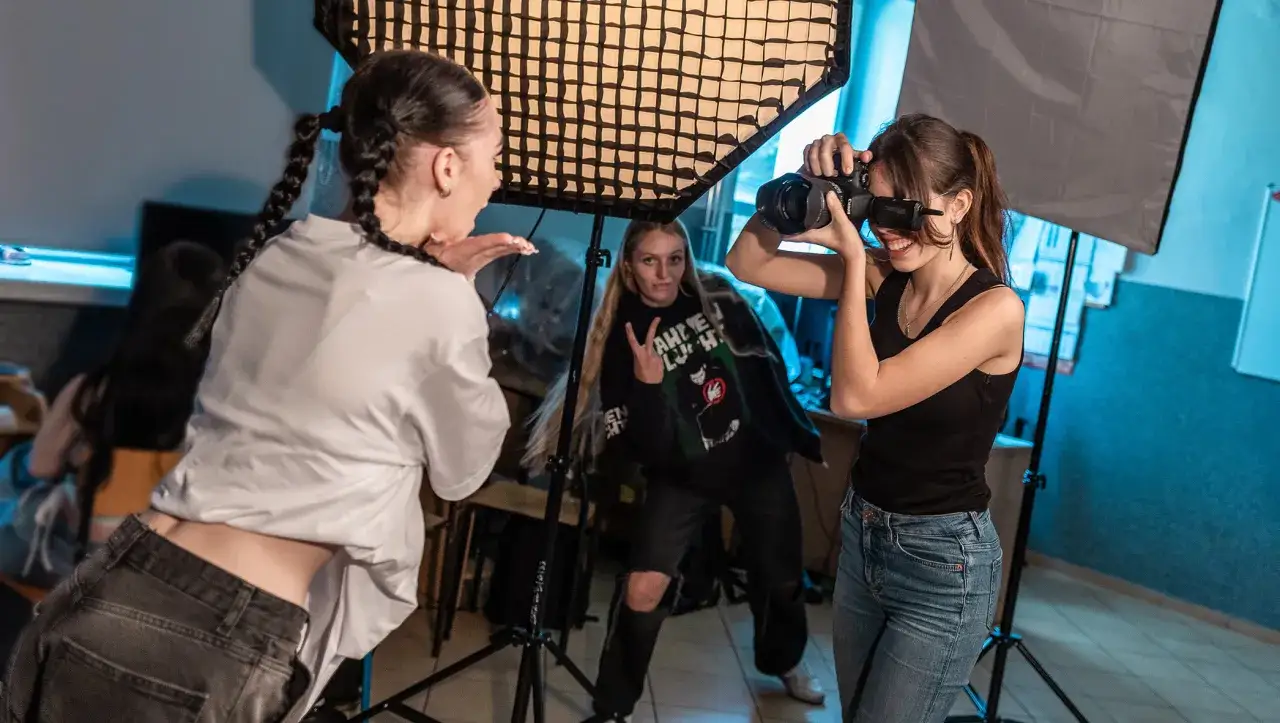 Uczennice branżowej szkoły 2 stopnia ćwiczą fotografię studyjną. Dziewczyna z aparatem robi zdjęcie koleżance pozującej pod lampą.
