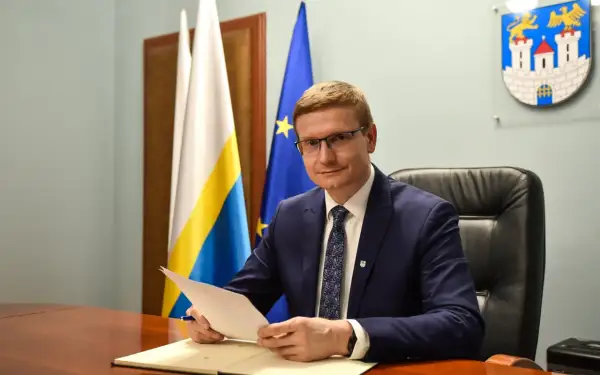 Mężczyzna w garniturze siedzi przy biurku, trzymając dokumenty. Za nim flagi i herb miasta, symbolizujące jego rolę w obszarze prawo administracyjne.