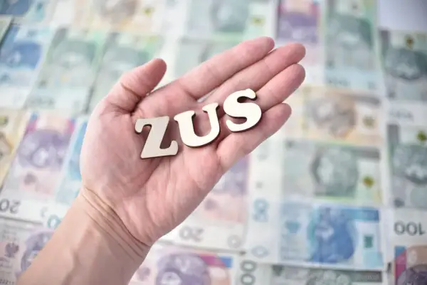 Drewniane litery ZUS leżą na dłoni, na tle polskich banknotów.