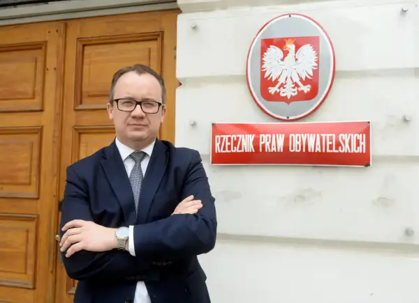 Mężczyzna w garniturze stoi przed budynkiem z godłem Polski i tabliczką "Rzecznik Praw Obywatelskich".
