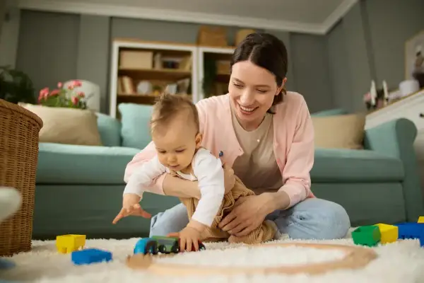Aktywni rodzice w pracy to nie tylko biuro. Uśmiechnięta mama bawi się z dzieckiem klockami i pociągiem na dywanie.